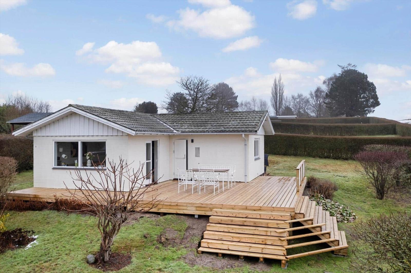 House with wooden terrace and garden. White facade, stairs, and seating area.