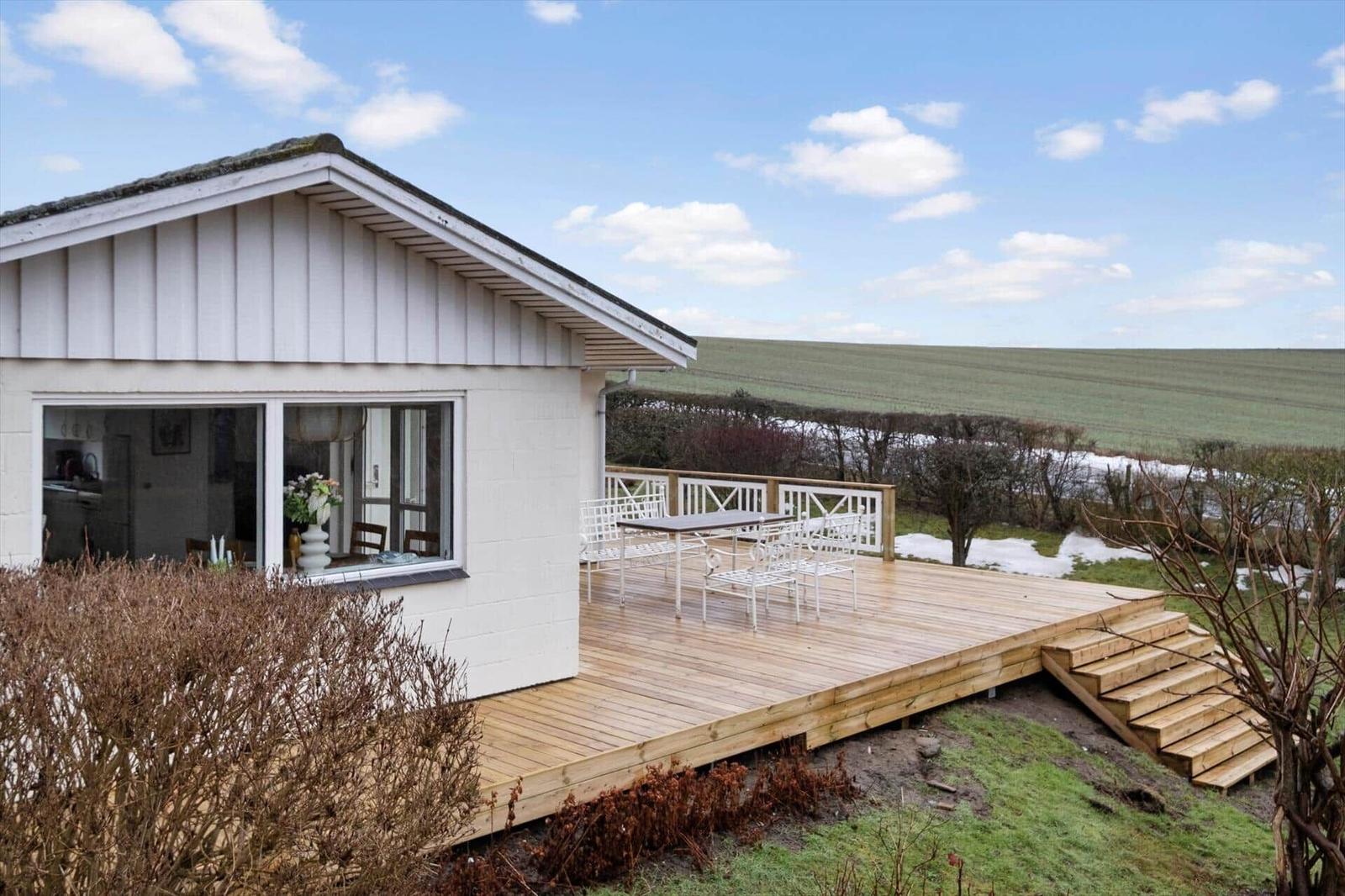 House with terrace, table, and chairs. View of fields and sky.