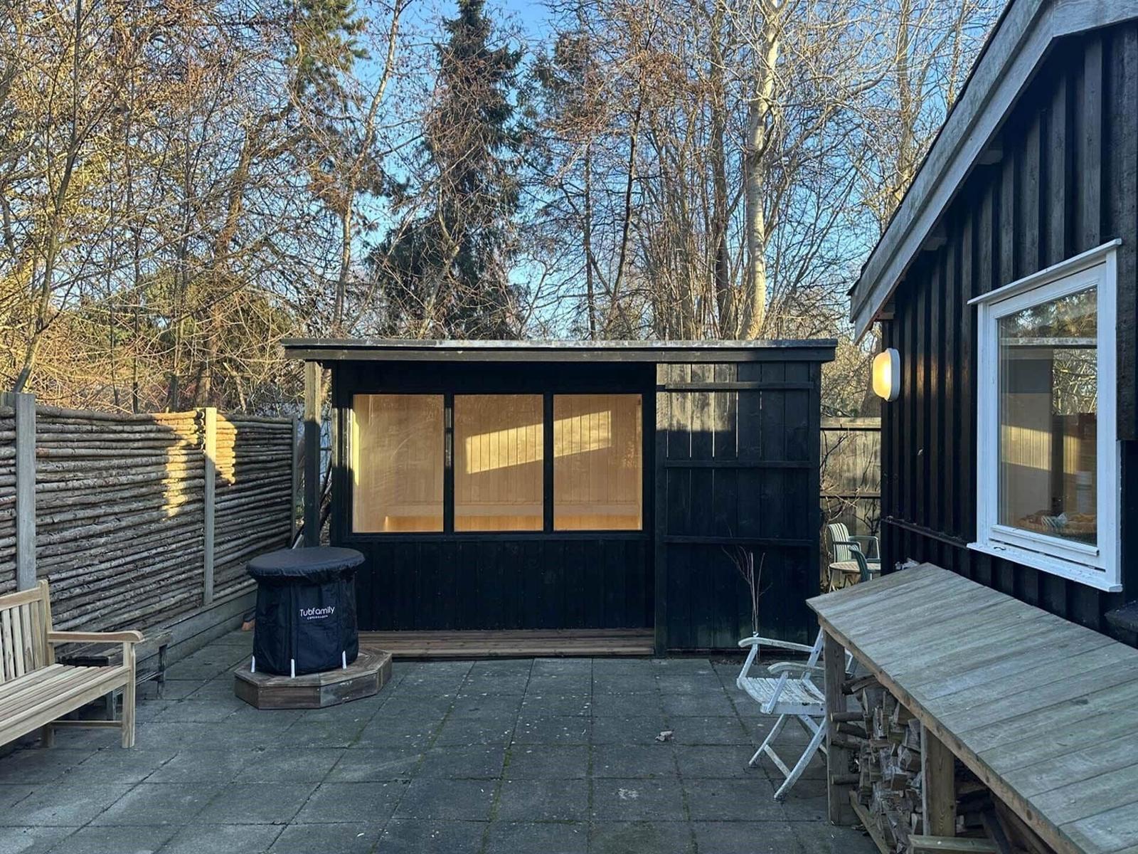 Backyard with sauna, wooden bench, folding chair, and woodpile. Surrounded by trees and log fence.