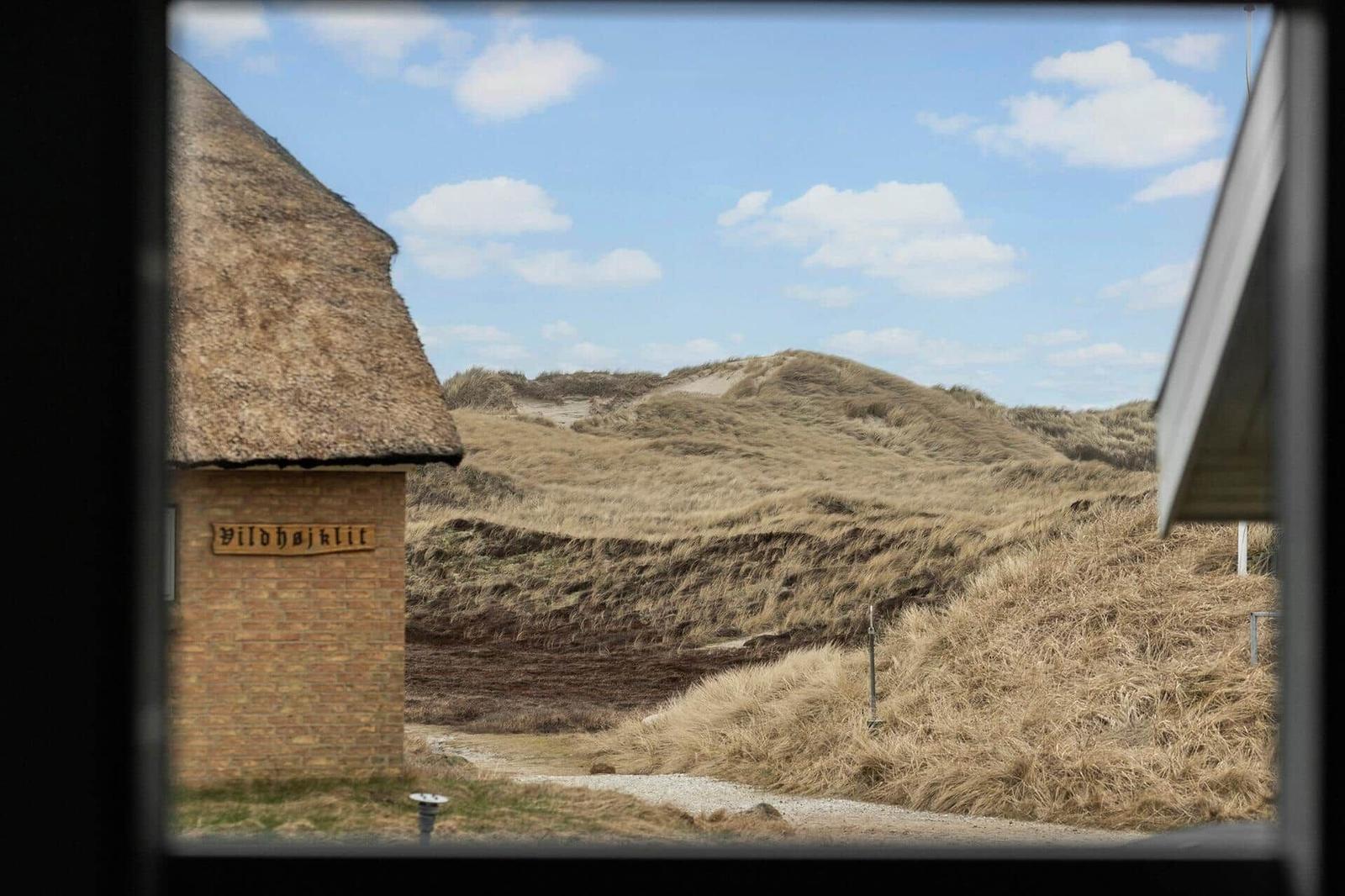Durch ein Fenster blickt man auf Dünenlandschaft mit einem Gebäude mit Strohdach und Schild.