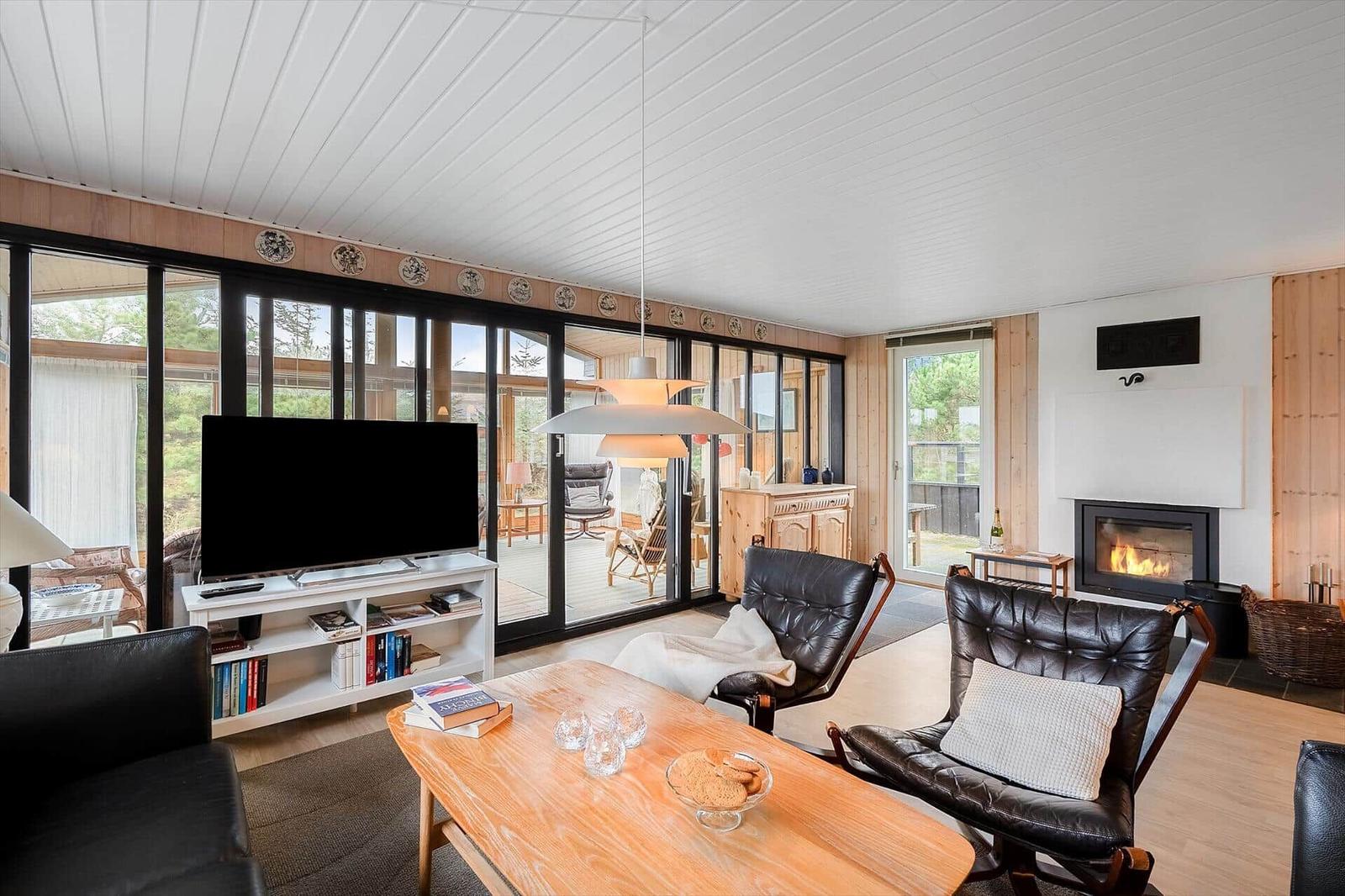Wohnzimmer mit Kamin, Fernseher und Sitzgelegenheiten. Große Glasfront zur Terrasse.