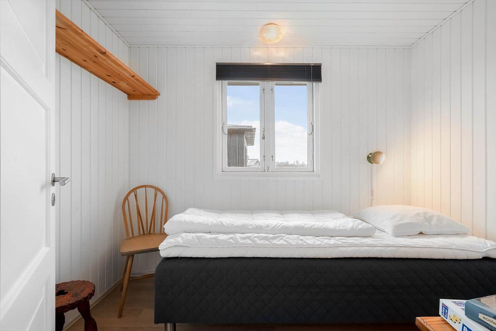 Schlafzimmer mit Bett, Stuhl und Fenster. Wände sind weiß verkleidet.