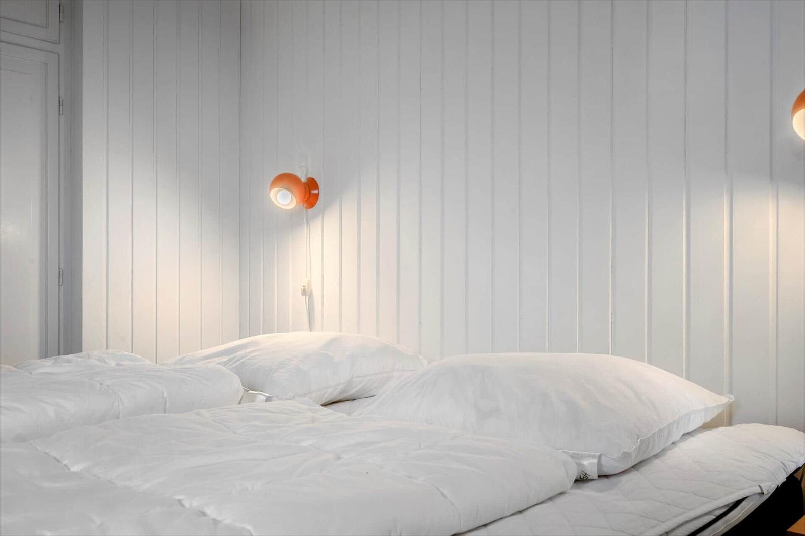 Schlafzimmer mit weißem Bett und orangefarbener Wandlampe an vertikal verkleideter Wand.