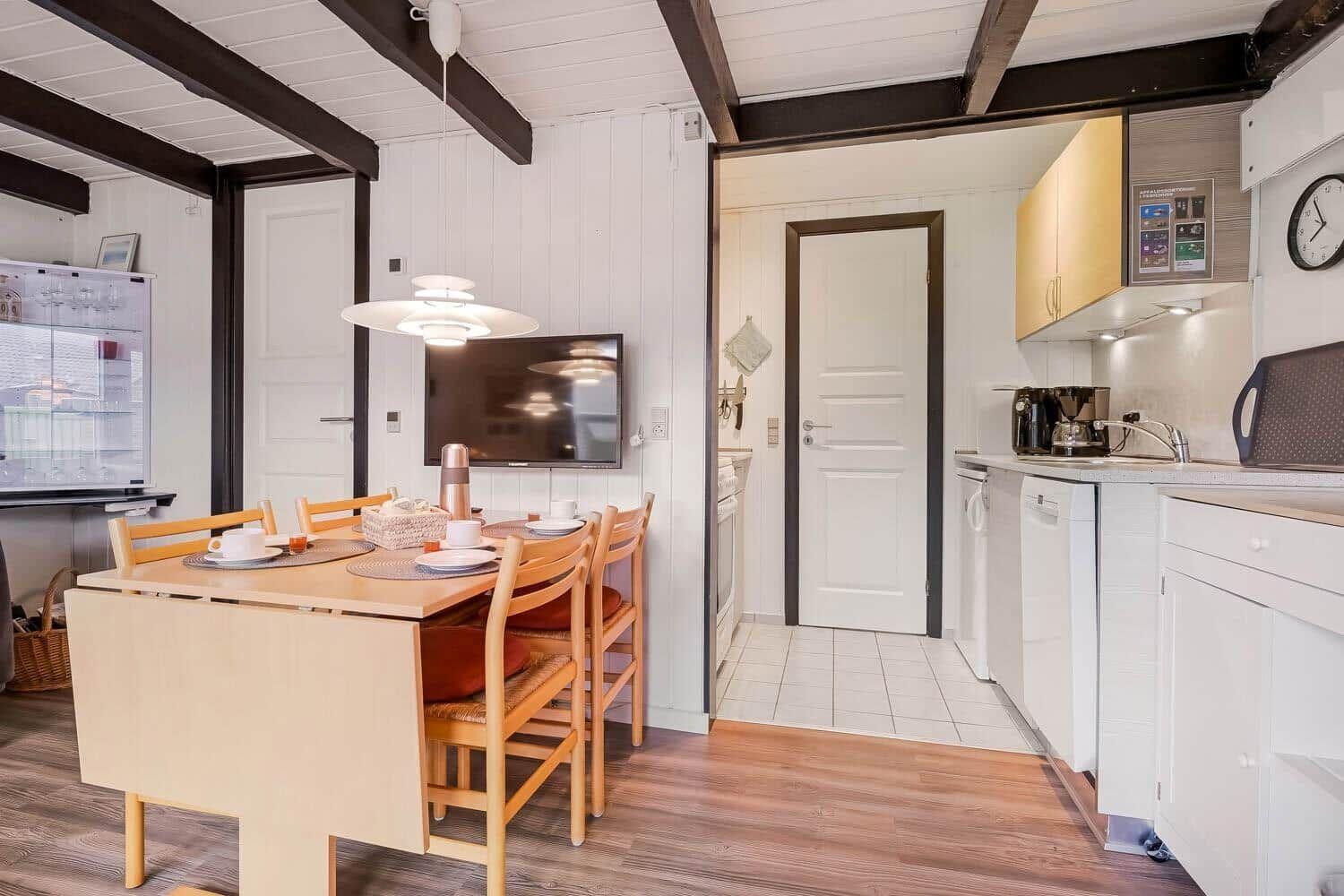 Essbereich mit Tisch und Stühlen, Küche mit Spüle und Kühlschrank, Fernseher an der Wand.