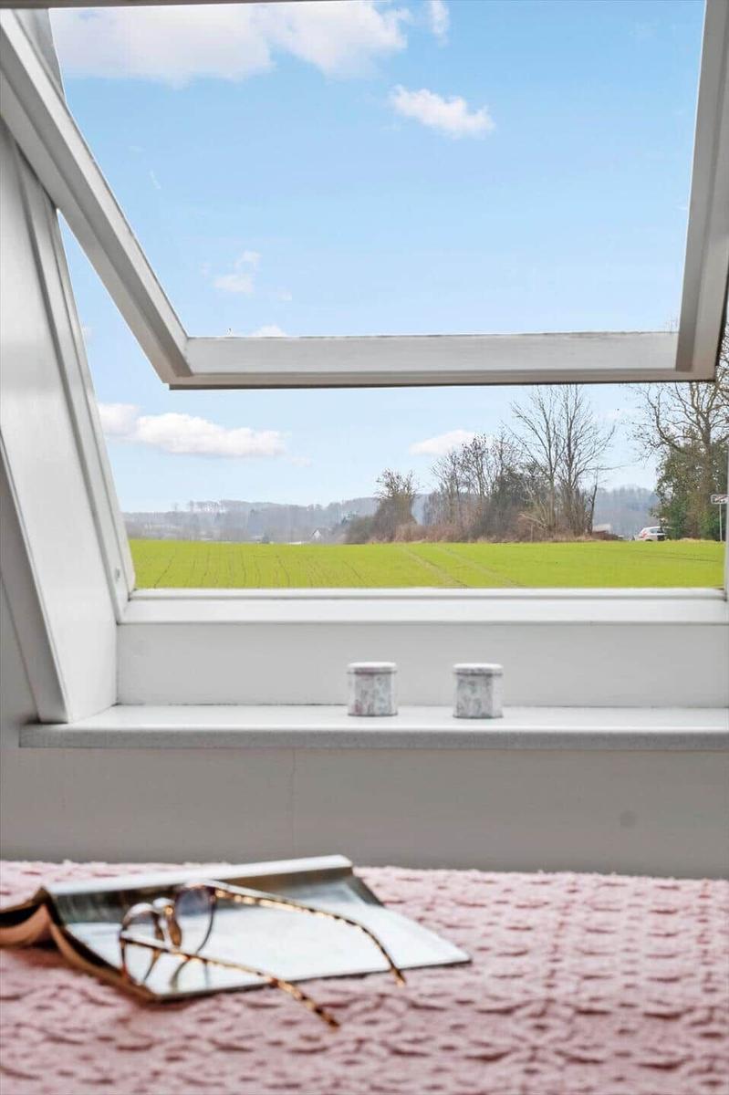 Durch das Fenster blickt man auf ein grünes Feld und einen blauen Himmel mit weißen Wolken.