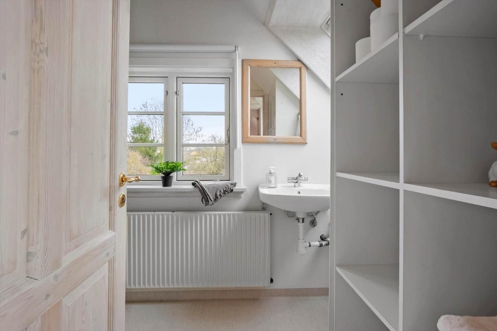 Badezimmer mit Waschbecken, Spiegel und Fenster. Regal und Heizung sind sichtbar.