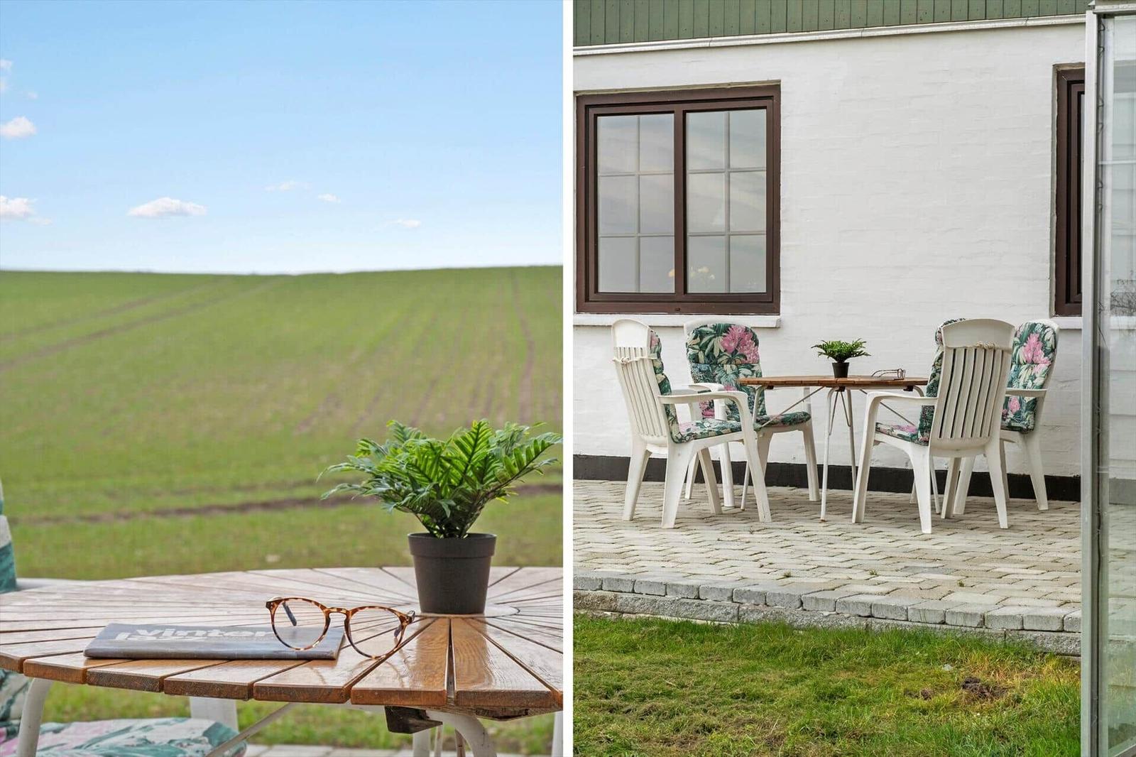 Terrasse mit Tisch und Stühlen, Blick auf grünes Feld und weißes Haus mit braunen Fenstern.