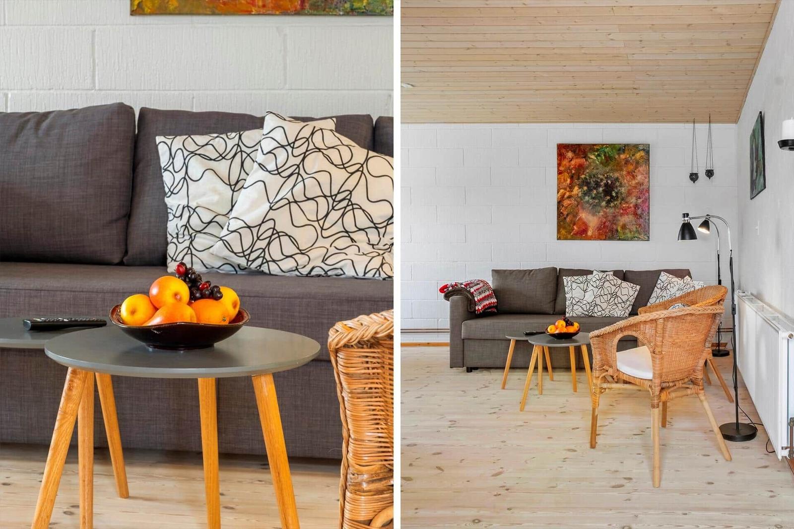 Wohnzimmer mit grauem Sofa, Holzdecke und Korbstühlen. Tisch mit Obst und Wandbild.