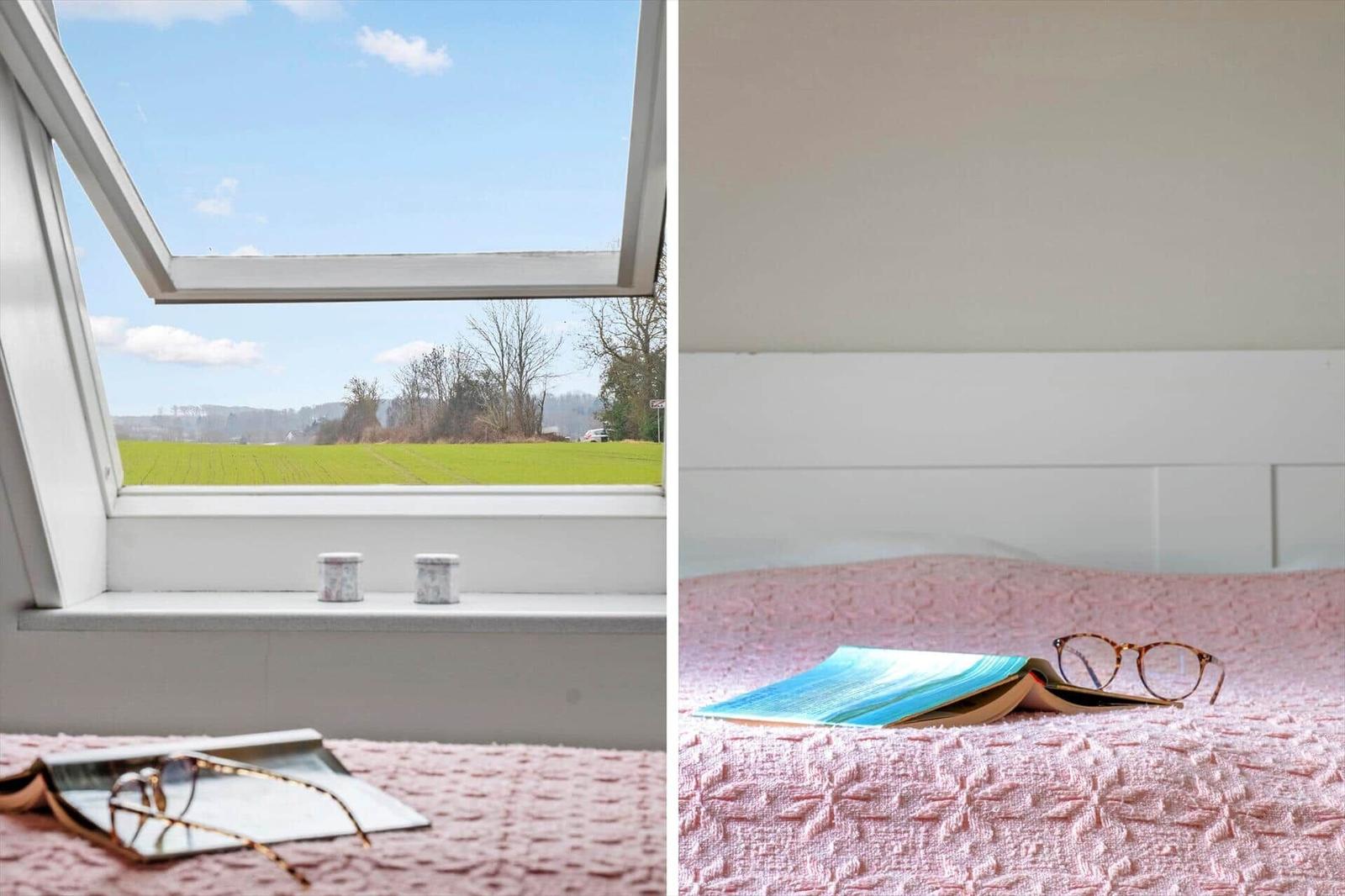 Dachfenster mit Blick auf Feld. Bett mit rosa Decke und Brille.