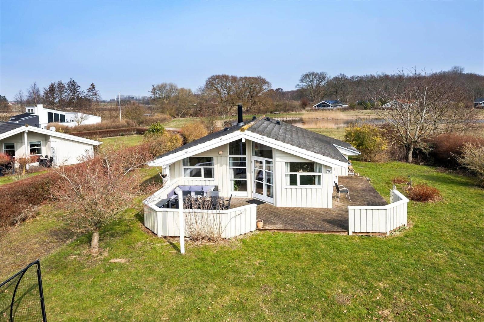 A white holiday home with a terrace and garden. Trees and a lake are visible in the background.