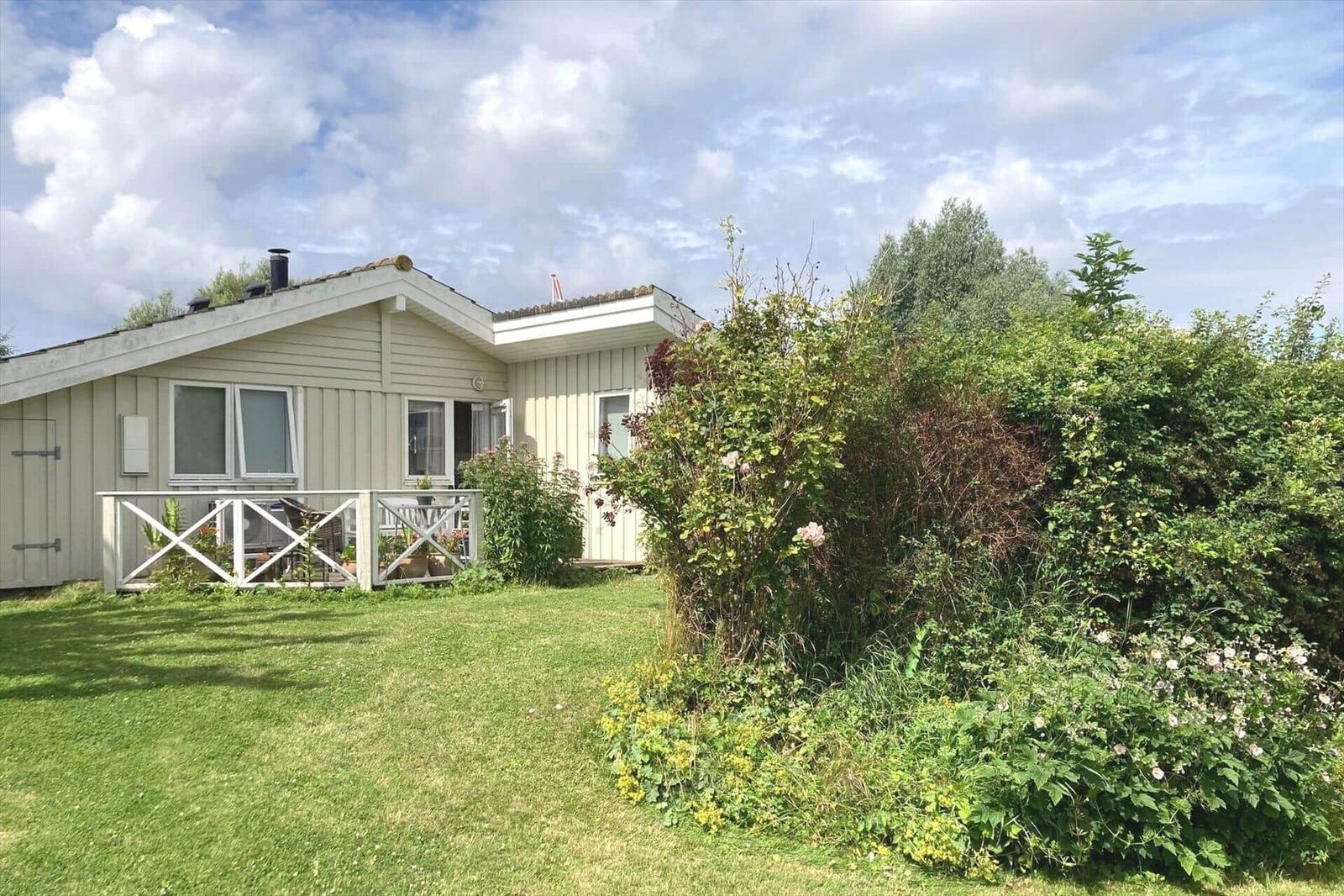 A small house with a terrace, surrounded by grass and bushes under a cloudy sky.