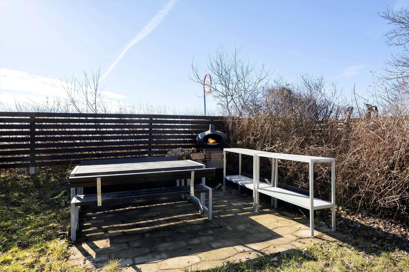 Outdoor area with table, benches, and wood stove. Background: fence and shrubs.
