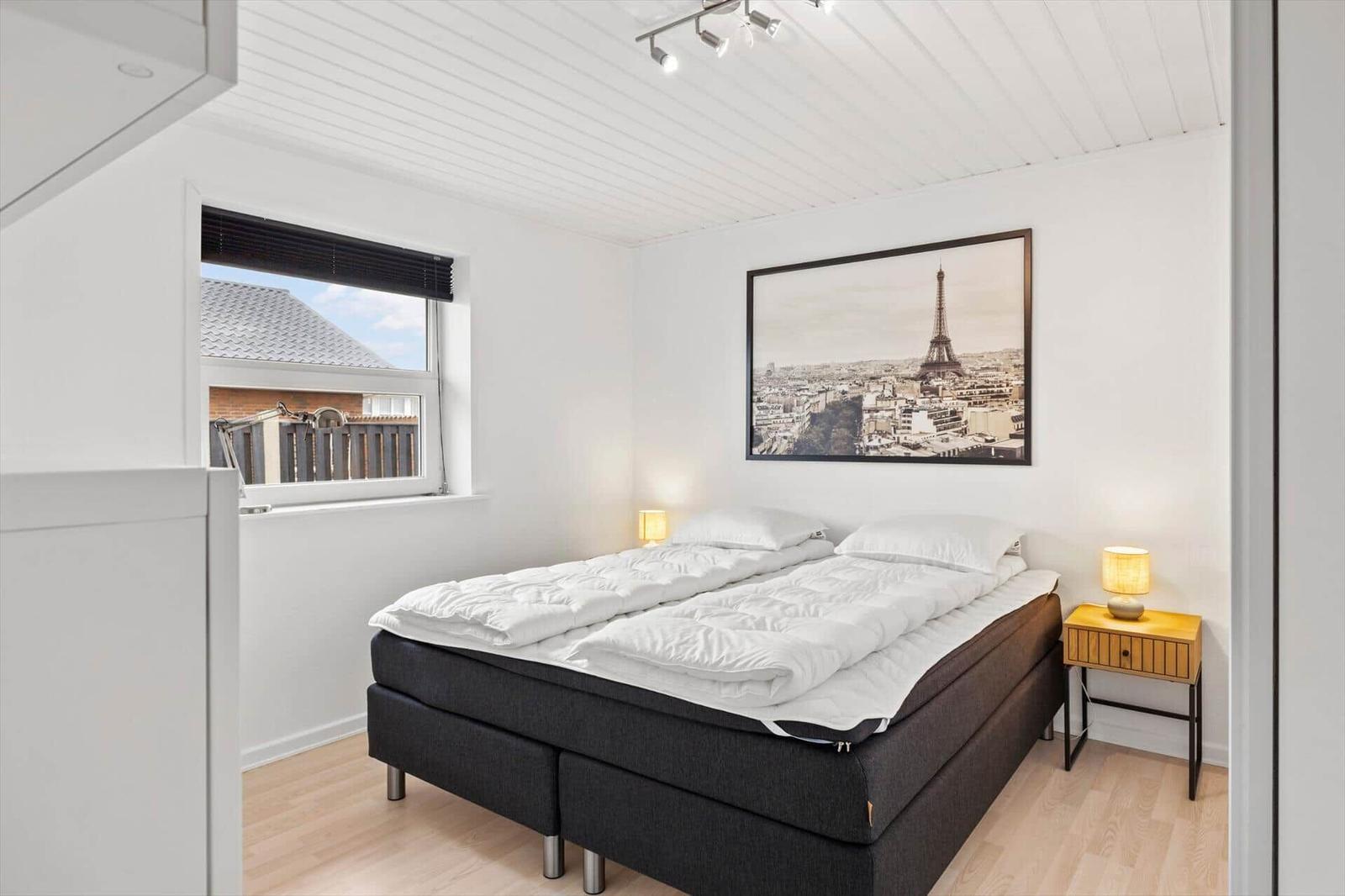 Schlafzimmer mit Bett, Nachttisch und Fenster mit Blick auf Dächer.