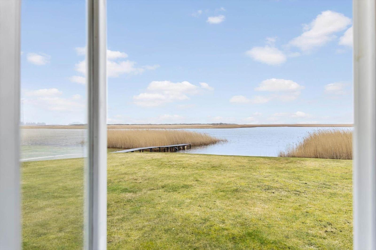 Blick aus dem Fenster auf einen See mit Ufergras und Steg.