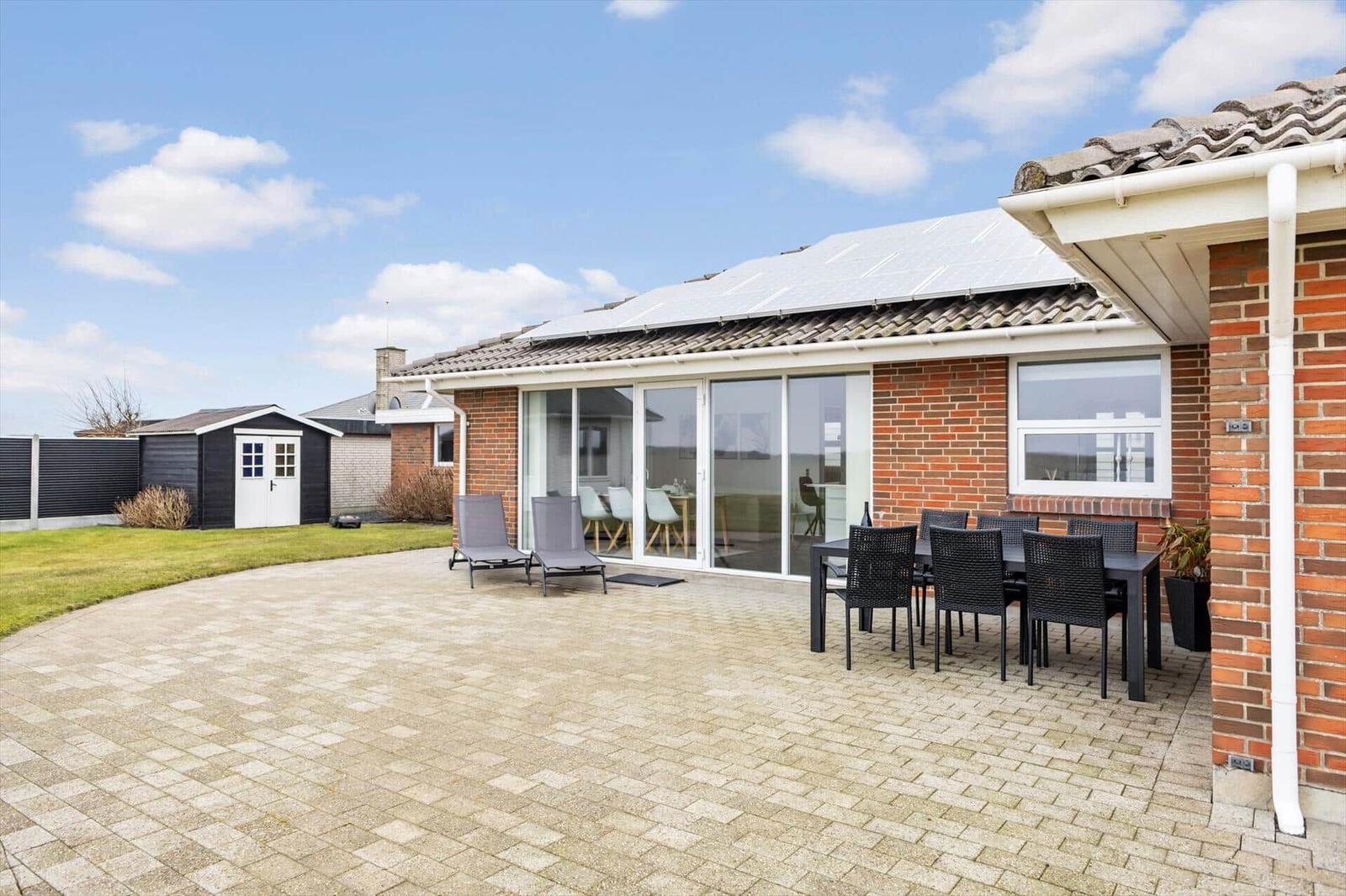 Terrasse mit Tisch, Stühlen und Liegestühlen. Glasfront zum Innenraum. Solarpaneelen auf Dach.