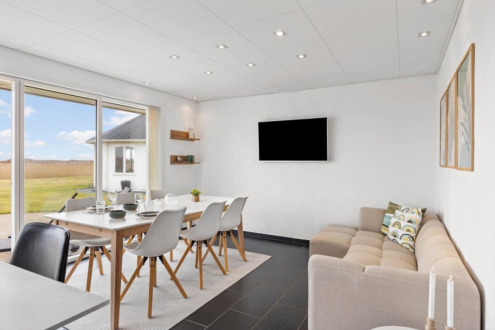 Wohn- und Essbereich mit Sofa, TV und Tisch mit Stühlen. Große Fensterfront mit Blick auf Grünfläche.