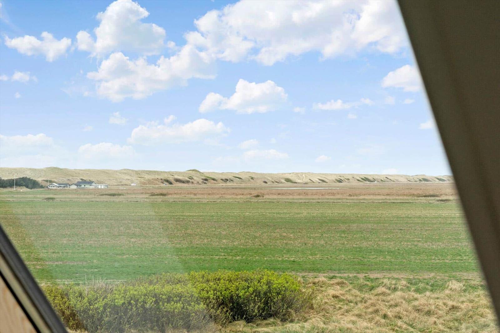 Weitläufiges Grünland mit Dünen im Hintergrund unter blauem Himmel mit weißen Wolken.