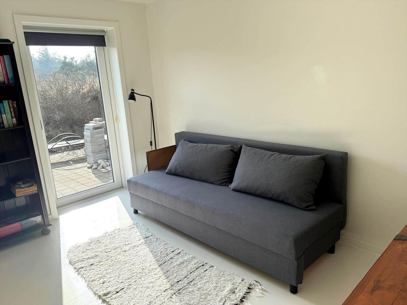 Ein grauer Sofa mit Kissen steht im Raum. Durch die Glastür ist ein Balkon sichtbar.