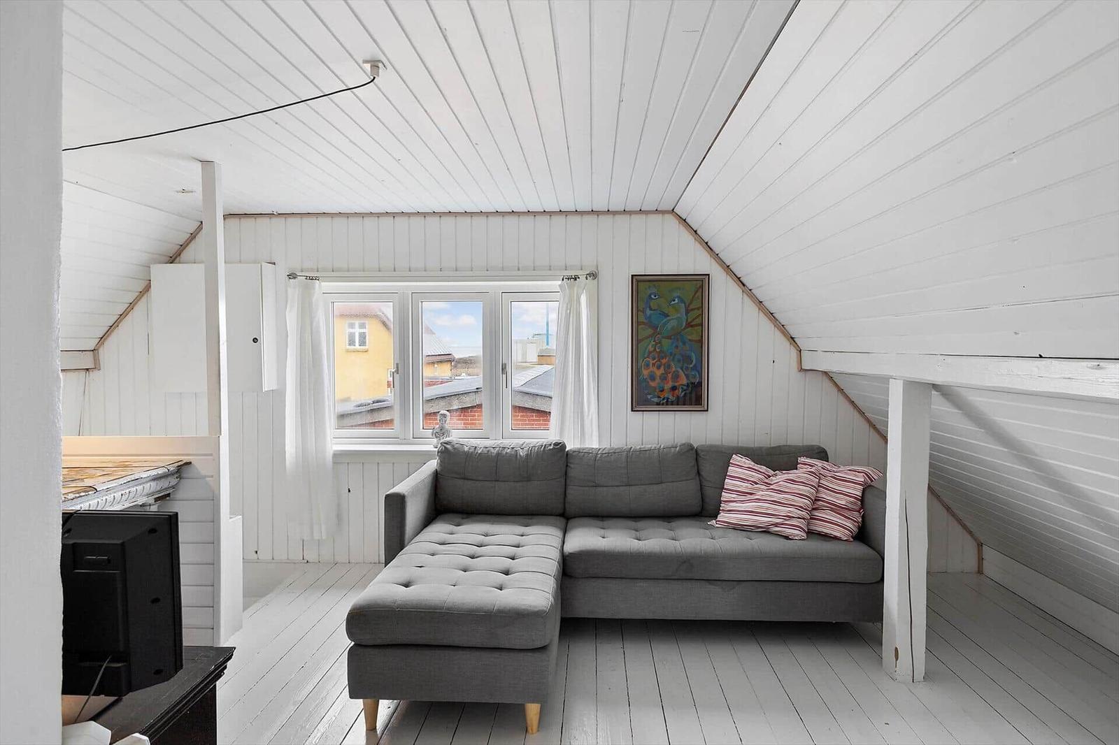 Wohnraum mit grauem Sofa, Fenster und Wandbild. Weiße Holzwände und Decke.