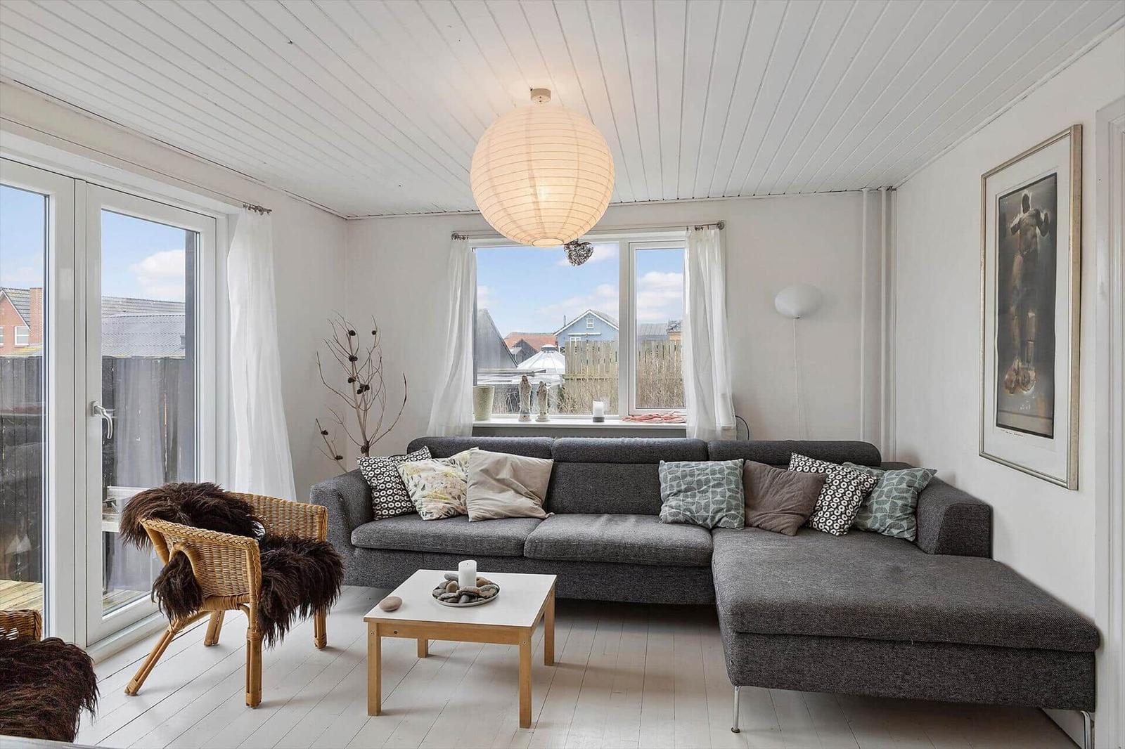 Ein Wohnzimmer mit grauem Sofa, Holztisch und Fenstern mit Blick nach draußen.
