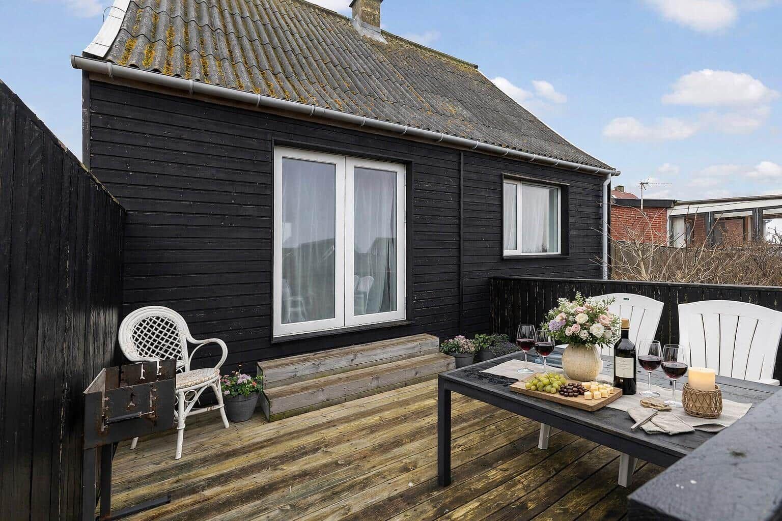 Terrasse mit Tisch, Stühlen und Weinarrangement vor einem schwarzen Haus.