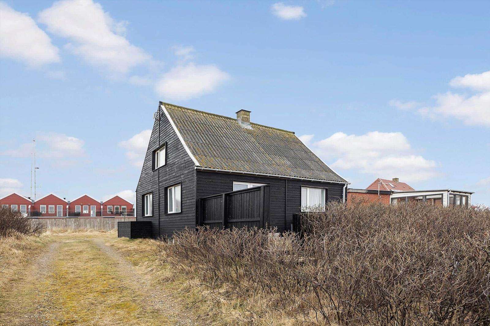Schwarzes Holzhaus mit Dach und Fenstern, umgeben von Sträuchern und einem Weg.
