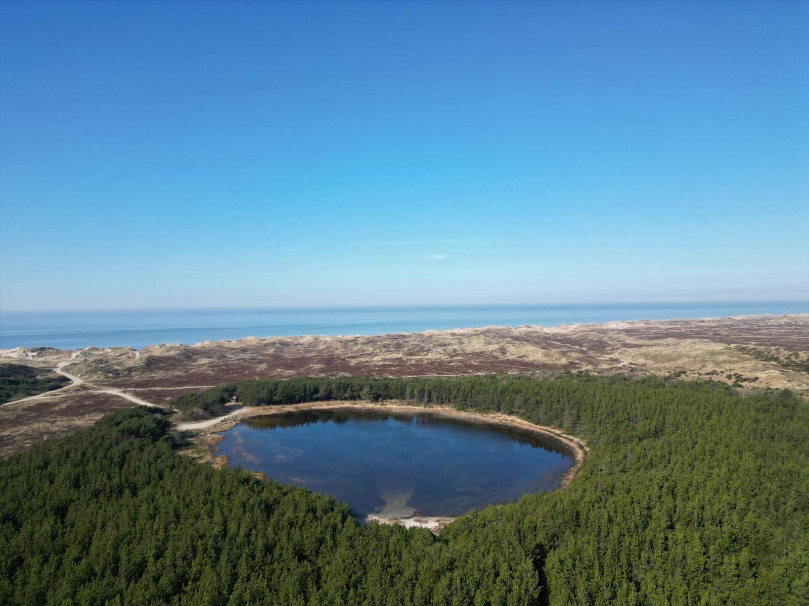 Ein See umgeben von Wald, mit Dünen und Meer im Hintergrund.
