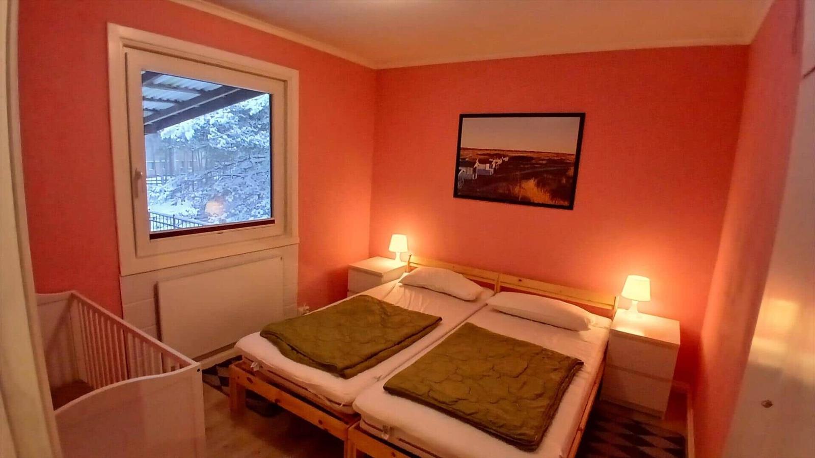 Zimmer mit Doppelbett, Fenster und Kinderbett. Wandfarbe rot. Beleuchtung durch Nachttische.