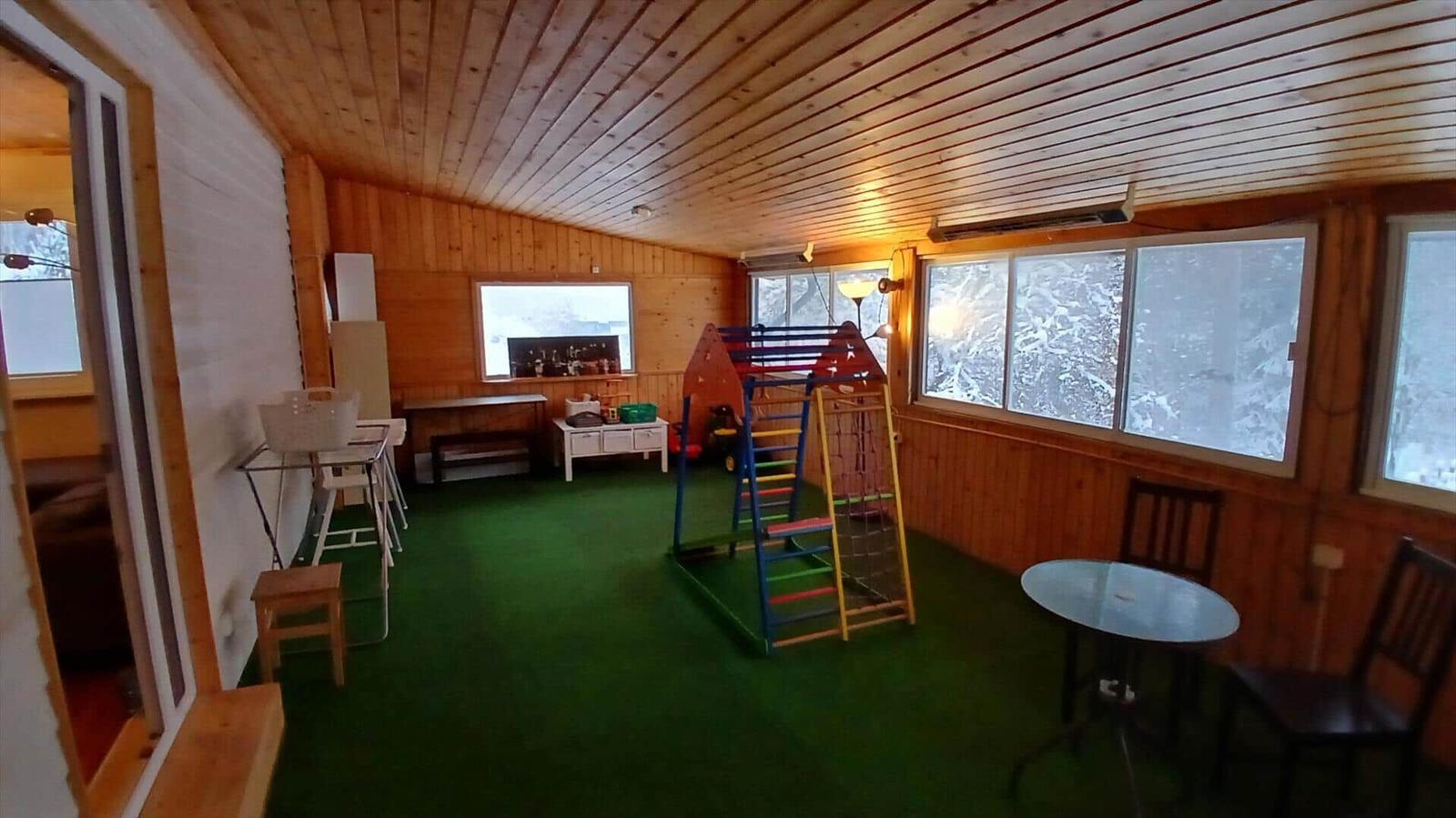 Räumlichkeiten mit Spielplatz, Tisch und Stühlen, grüner Boden, Holzwände und Fenster mit Schneeansicht.