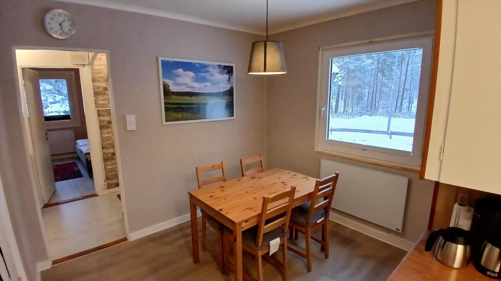 Esszimmer mit Holztisch, Stühlen und Fensterblick auf Wald. Küche und Schlafzimmer sichtbar.
