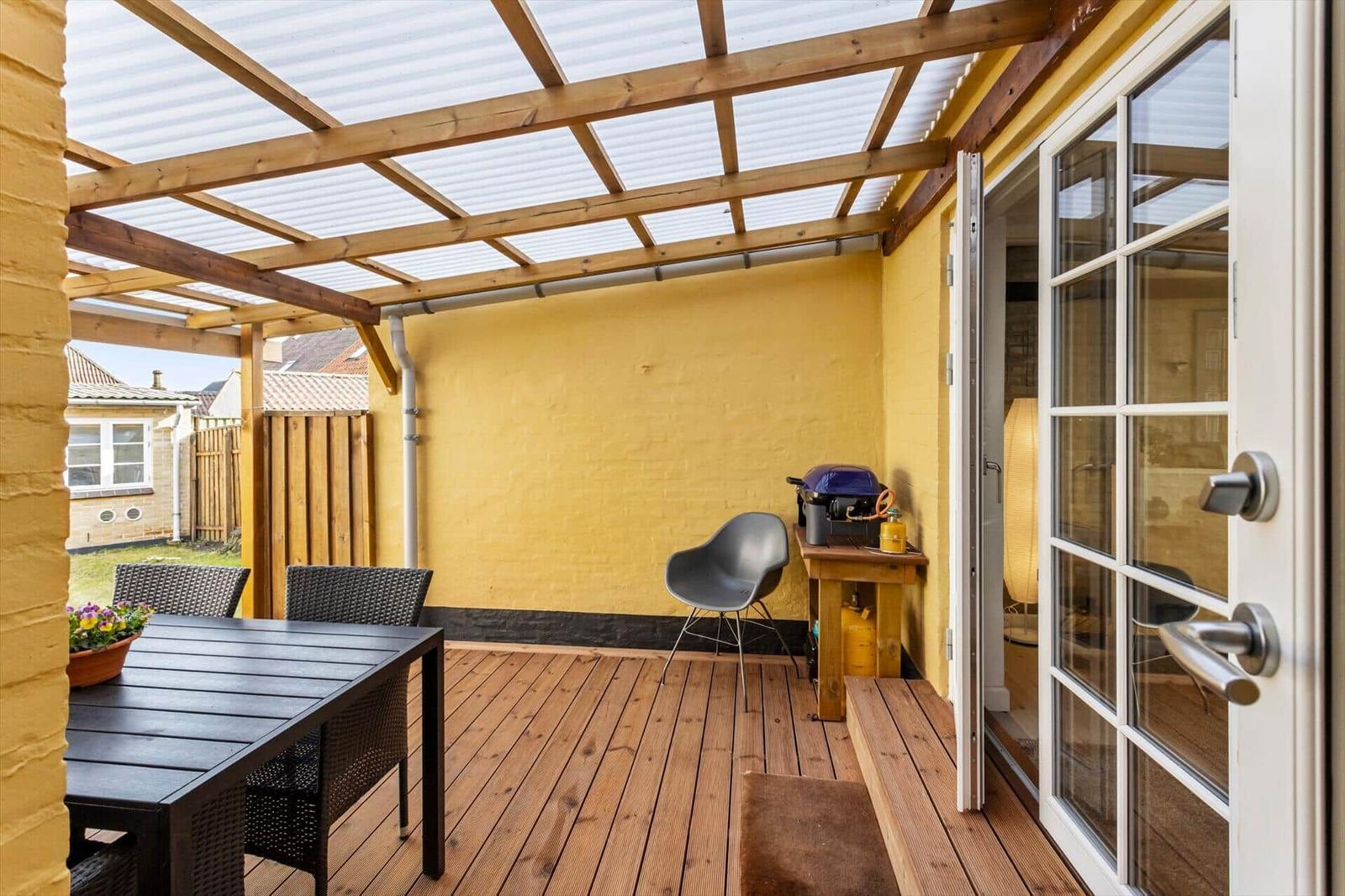 Deck with wooden roof, table, chairs, and grill. Glass door leads to the apartment.