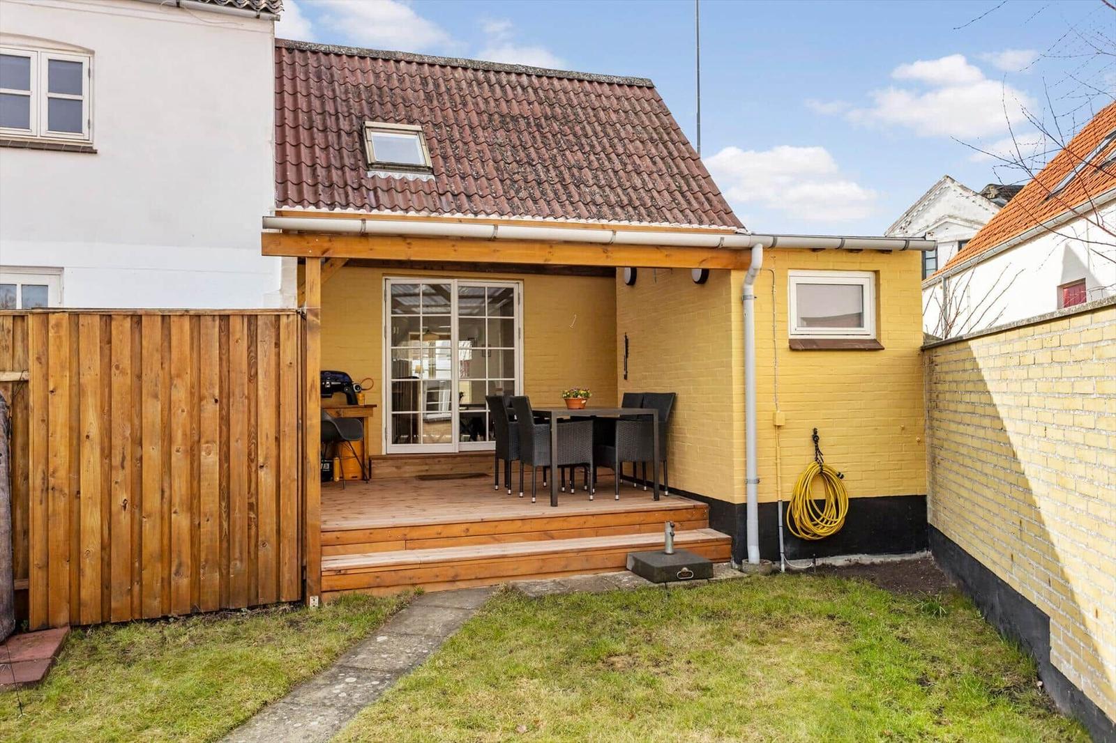 Backyard with terrace, table, chairs, and grill. Yellow extension with roof and window.
