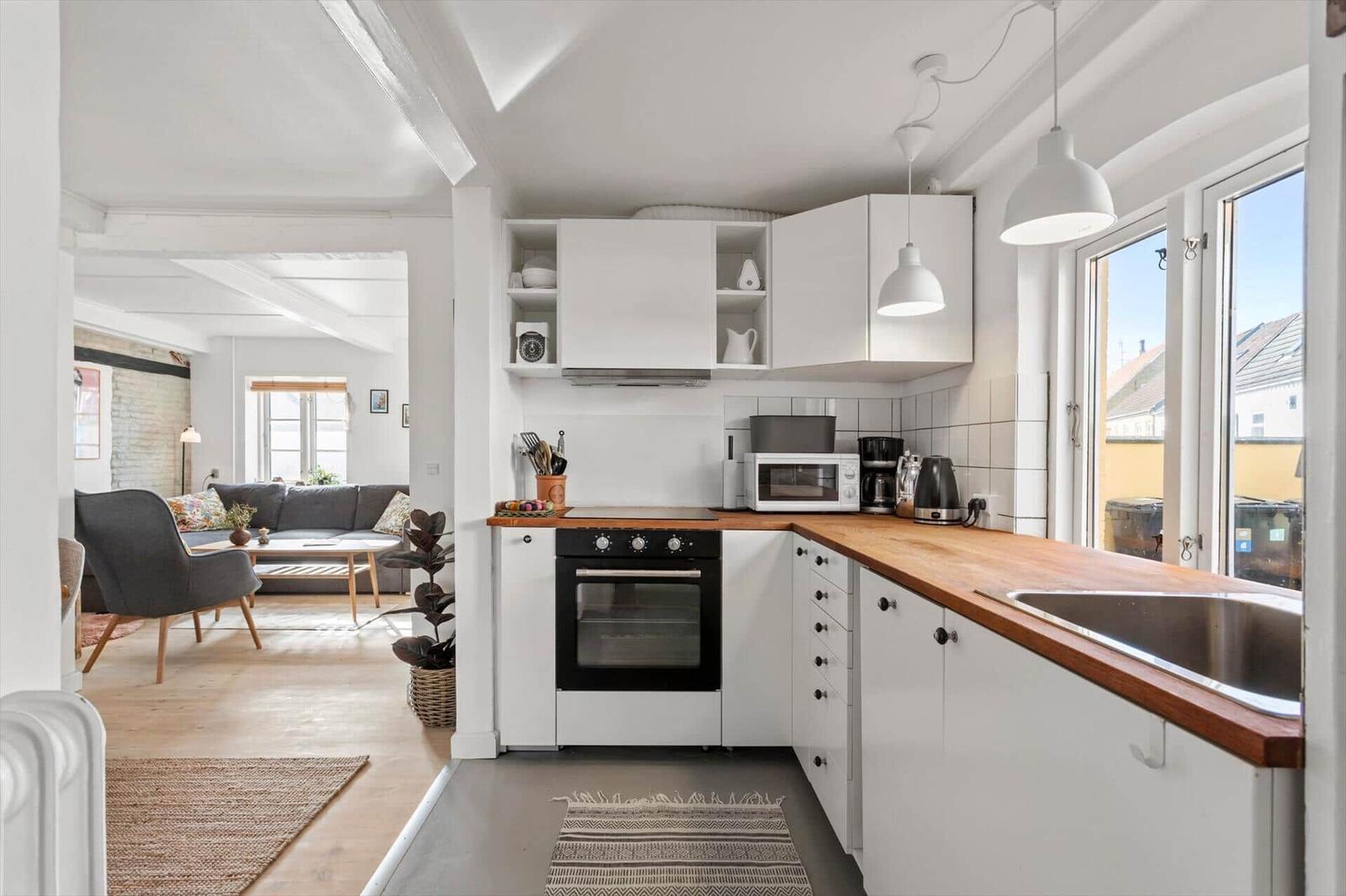Kitchen with white cabinets and wooden countertop. Living room with sofa in background.