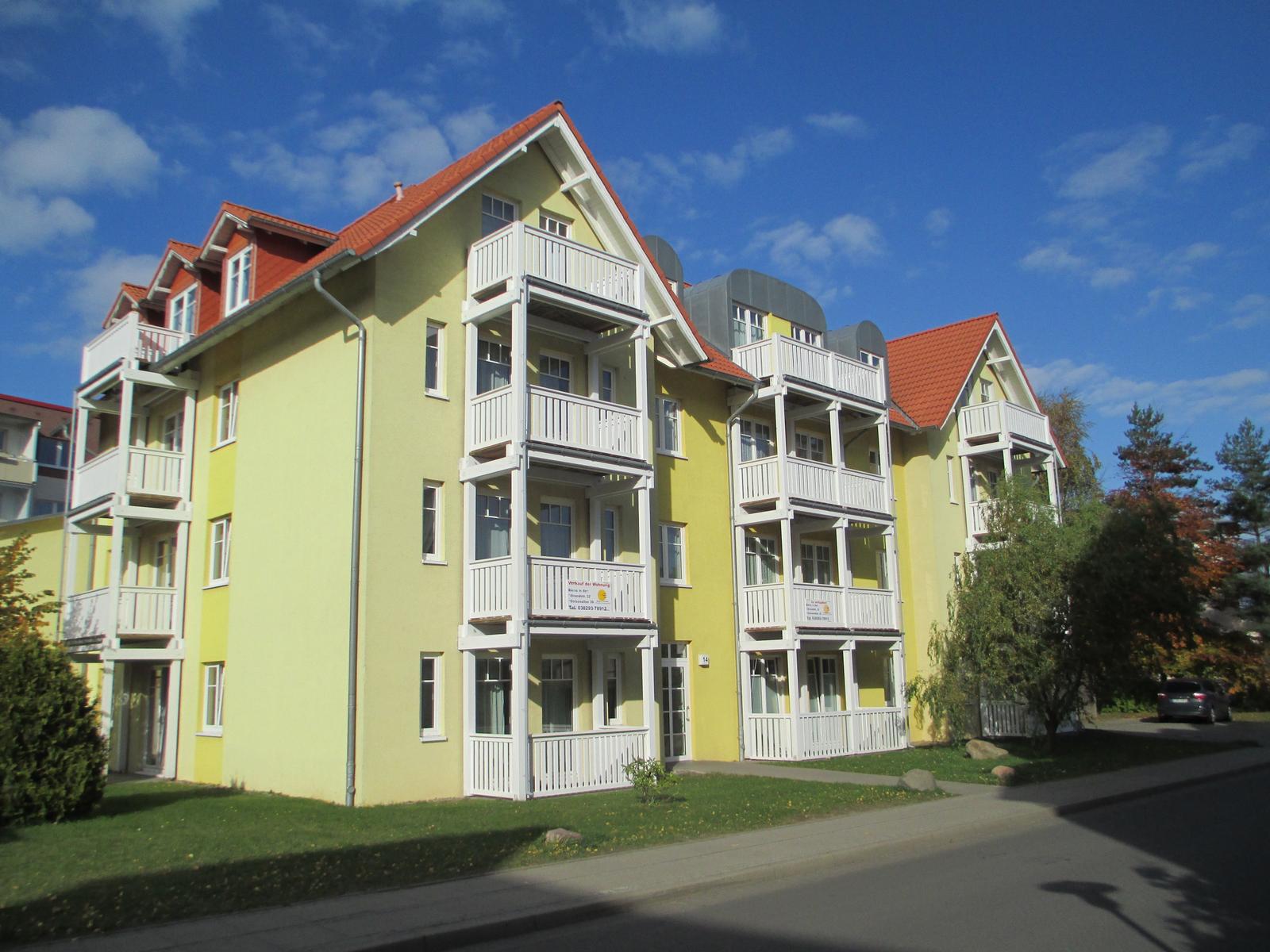 Gelbes Mehrfamilienhaus mit weißen Balkonen und rotem Dach unter blauem Himmel.
