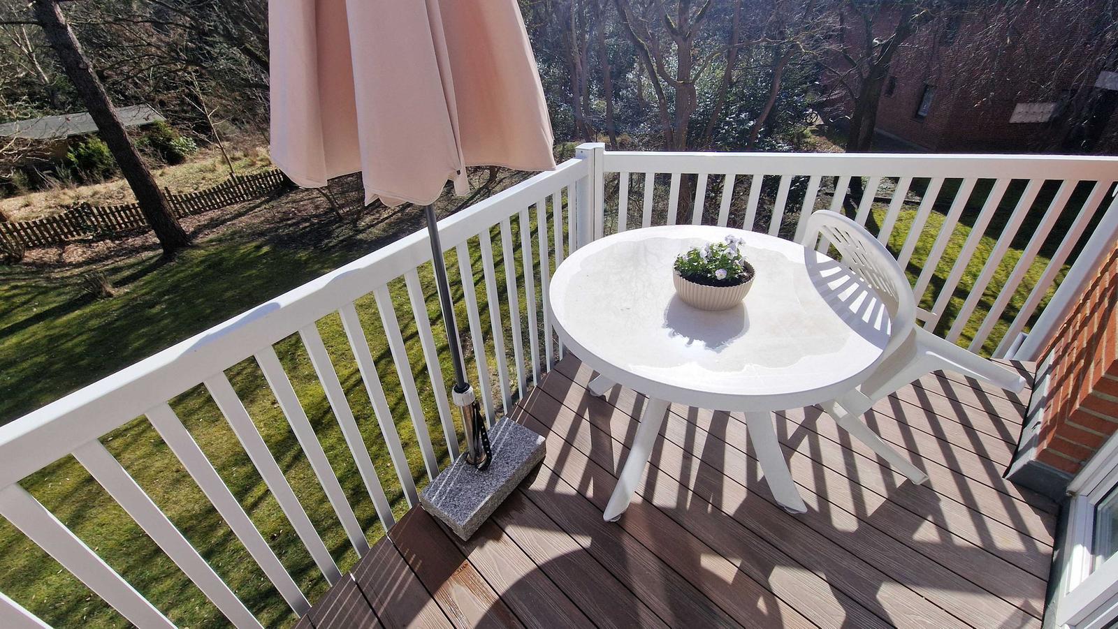 Terrasse mit weißem Tisch, Stuhl und Sonnenschirm. Blick auf grünen Garten und Bäume.