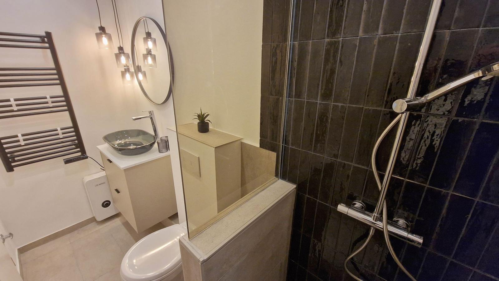 Bad mit Dusche, Waschbecken und Toilette. Schwarze Fliesen und moderne Beleuchtung.