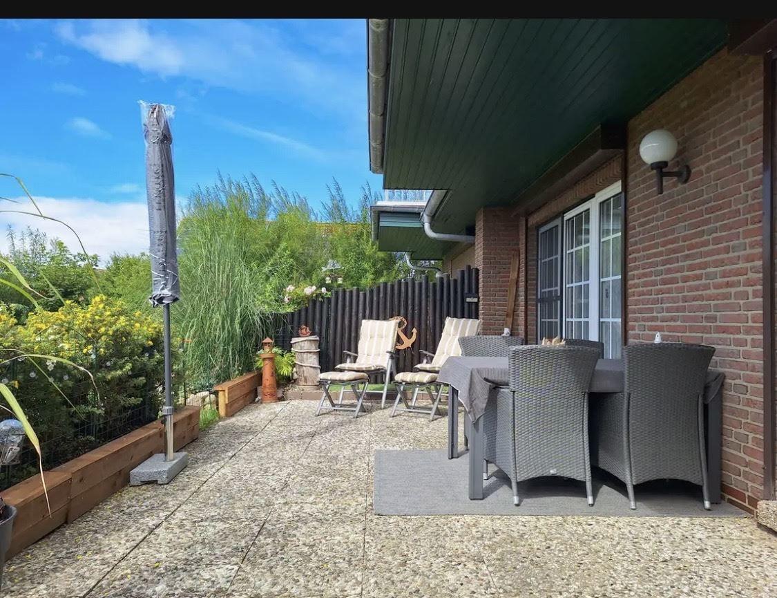 Terrasse mit Tisch, Stühlen und Sonnenschirm. Hintergrund: grüne Pflanzen und Zäunung.