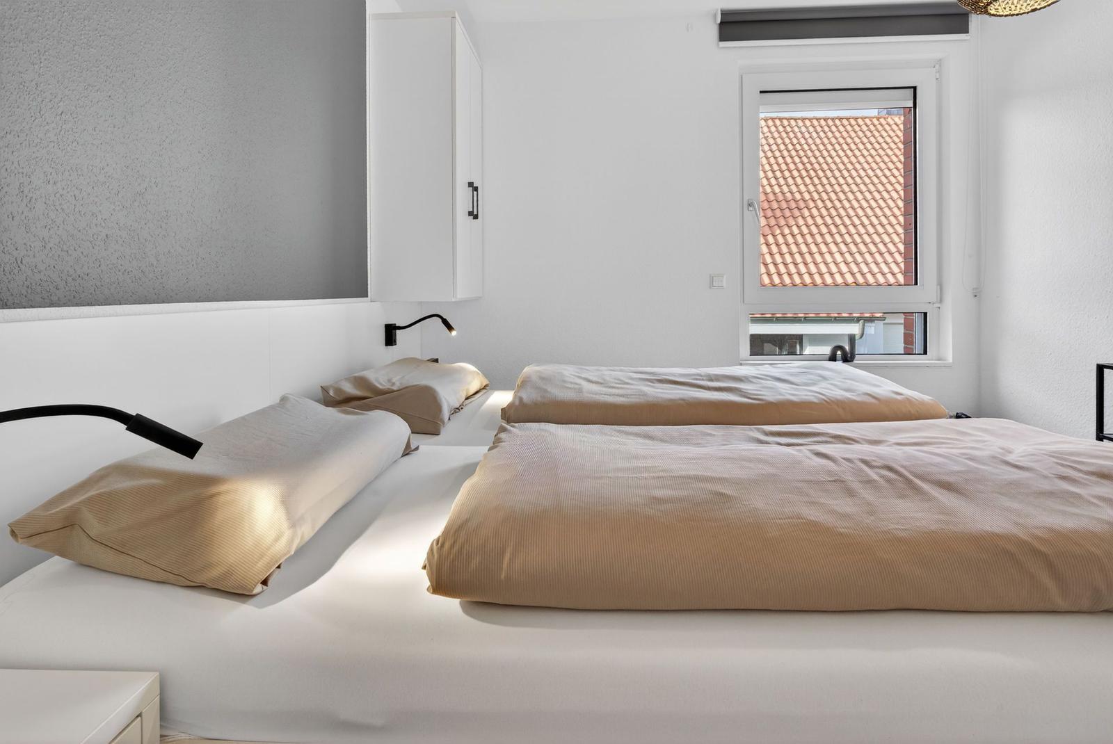 Zwei Betten mit hellen Bettwäsche, Fenster mit Dachziegeln und Schrank an der Wand.