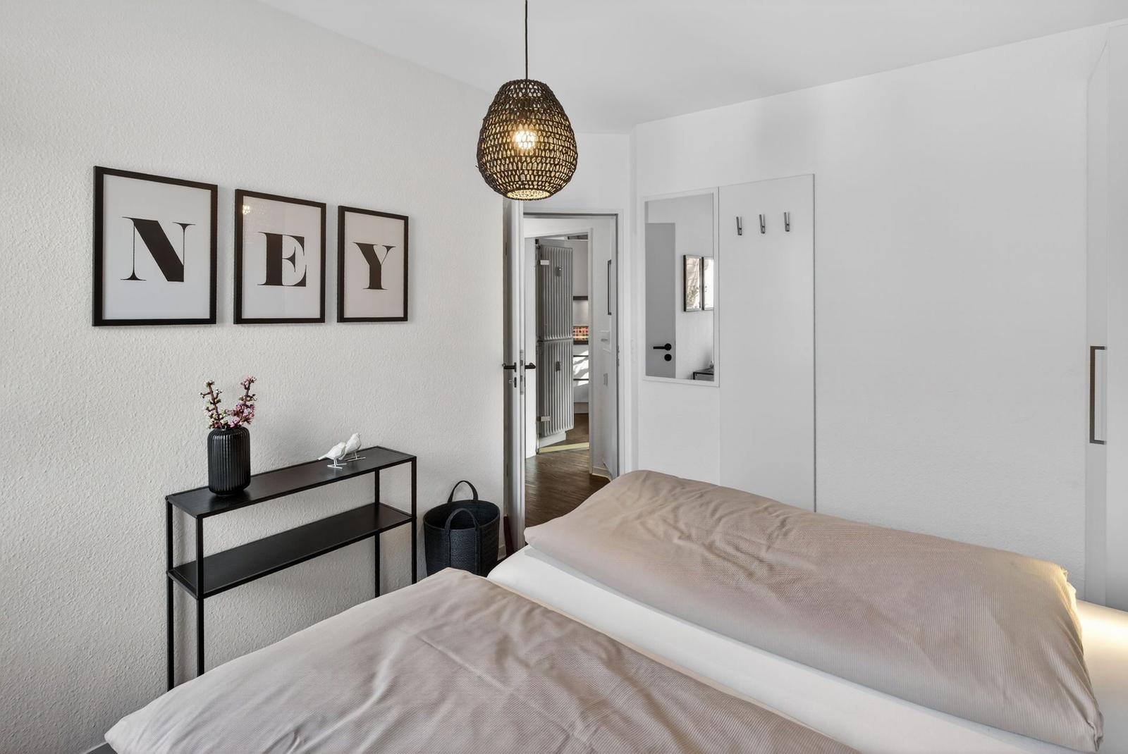 Schlafzimmer mit Bett, Wandbildern und Deckenleuchte. Blick in angrenzenden Raum.