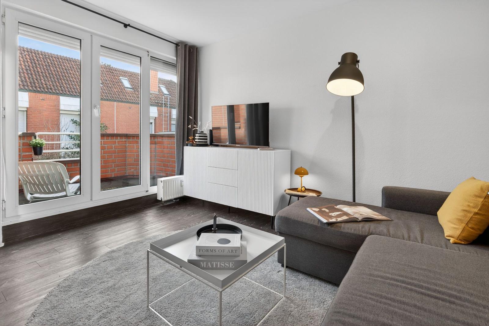 Wohnzimmer mit Sofa, Couchtisch und TV. Durchgang zu Balkon mit Sitzgelegenheit.