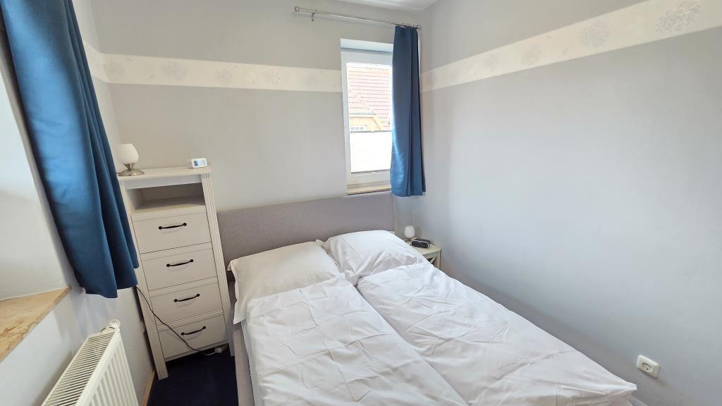 Schlafzimmer mit Doppelbett, Nachttisch, Schrank und Fenster mit blauen Vorhängen.