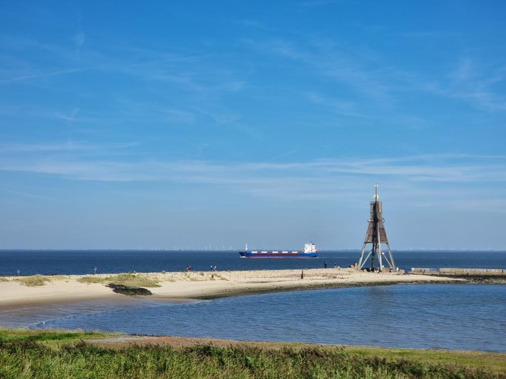 Hafen mit Segelboot, Sandstrand und Windkraftanlagen im Hintergrund.