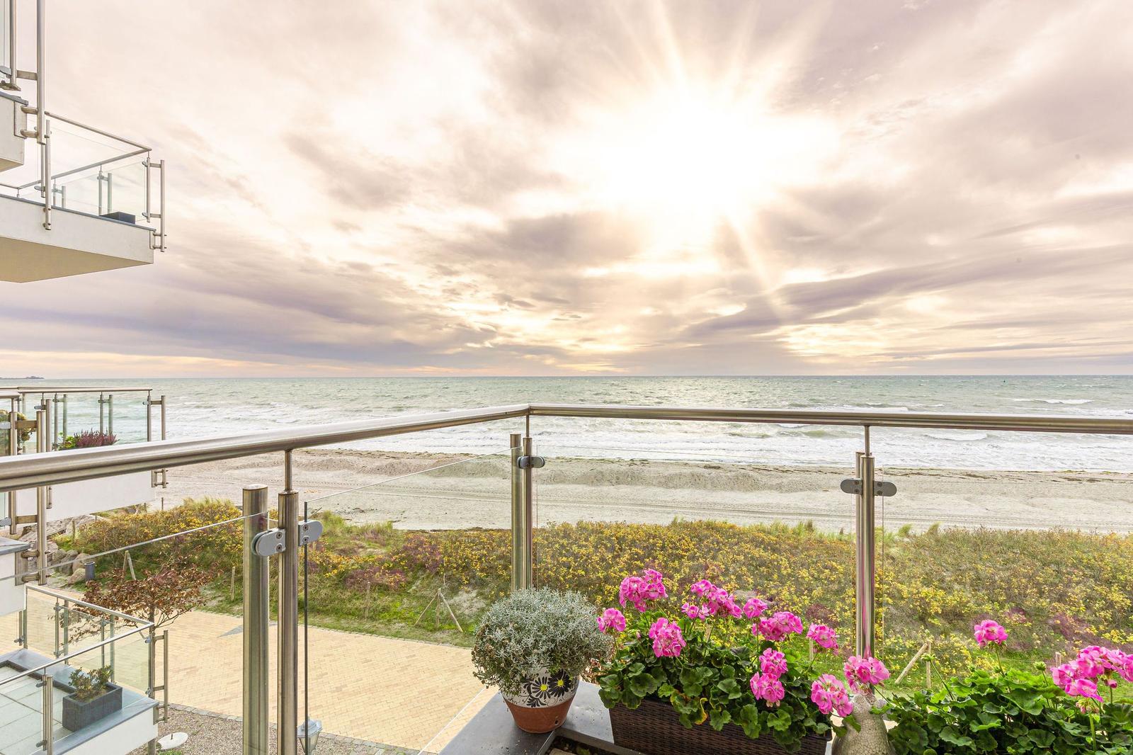Terrasse mit Blick auf Strand und Meer bei Sonnenuntergang. Blumen und Glasgeländer sind sichtbar.