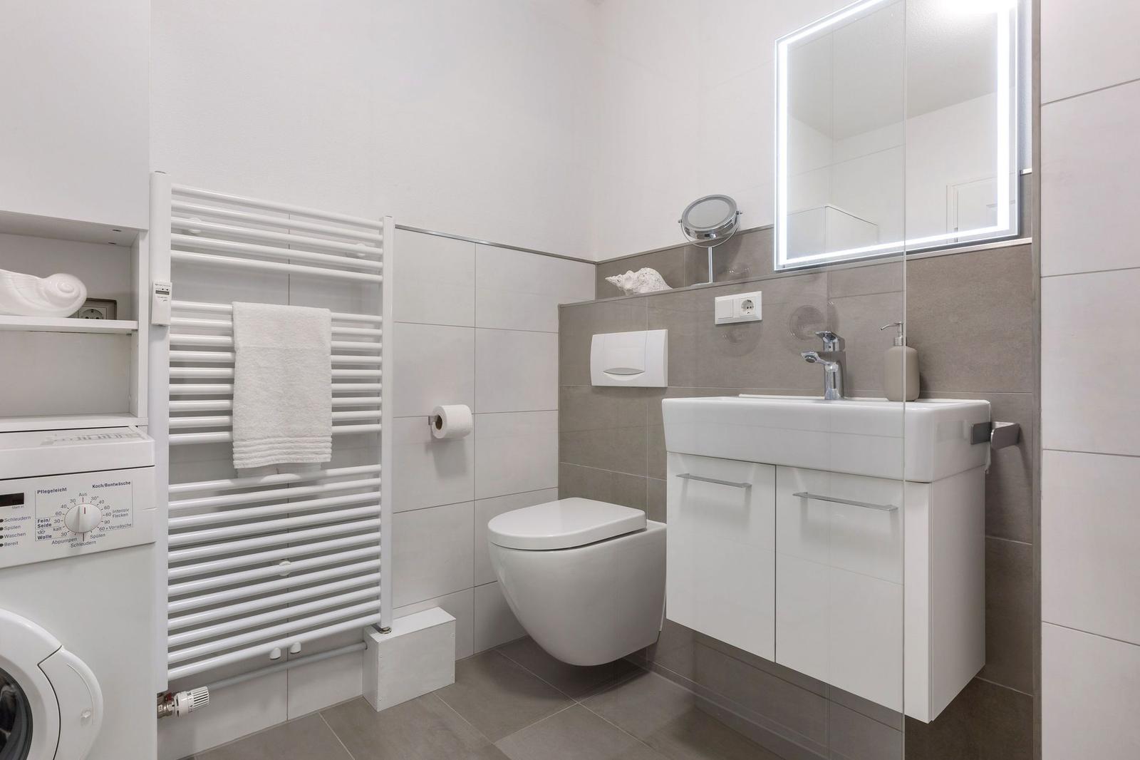 Badezimmer mit Toilette, Waschbecken und Heizung. Weiße Fliesen und graue Wandfliesen.