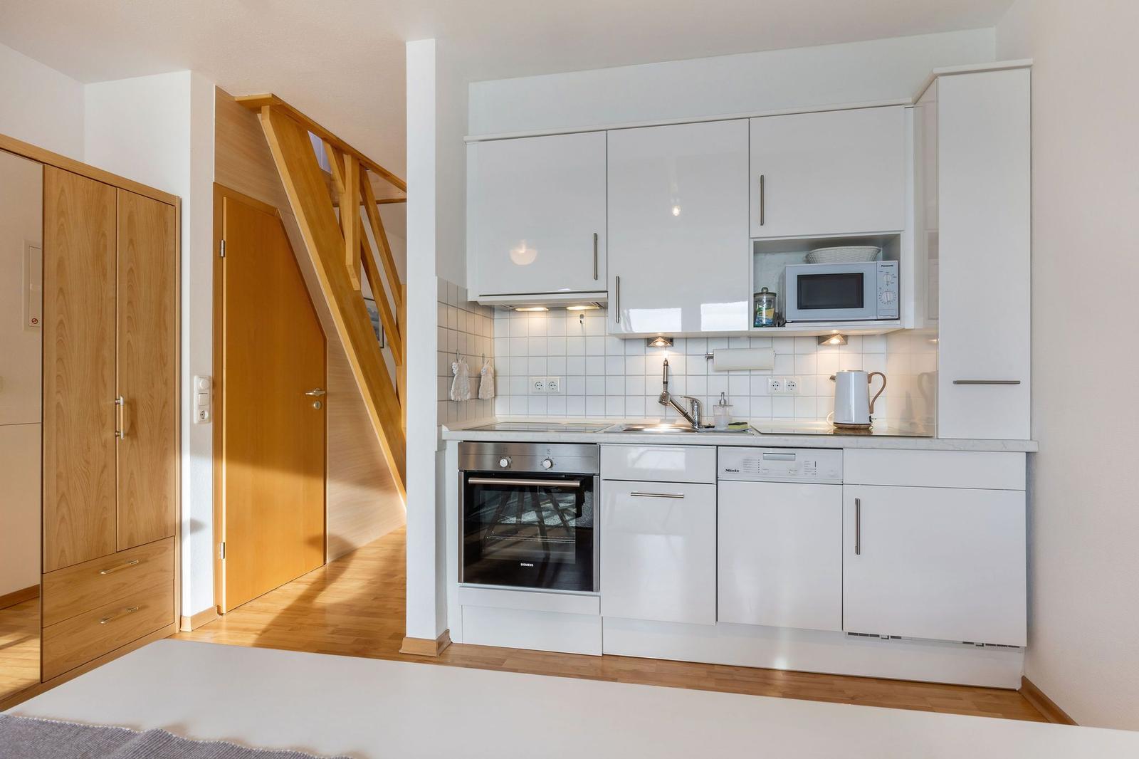 Küche mit weißen Schränken, Spüle, Herd und Mikrowelle. Holztreppe und Schrank im Hintergrund.