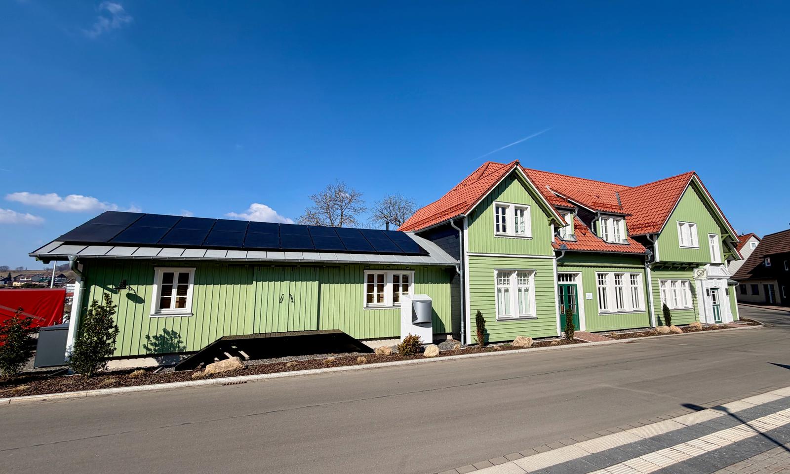 Grünes Haus mit Solaranlage und rotem Dach. Straße und kleine Bäume sind sichtbar.