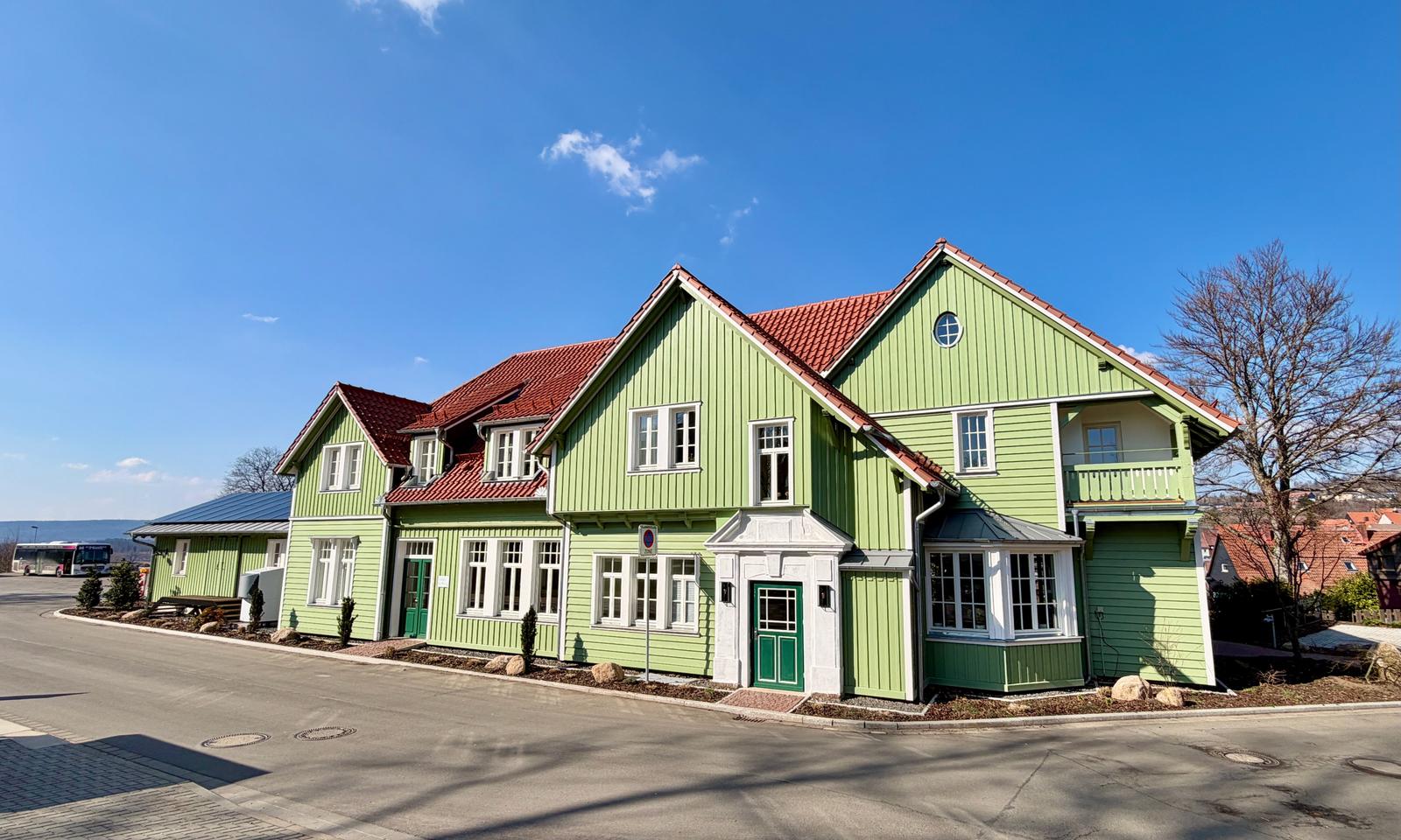 Grünes Holzhaus mit rotem Dach und weißen Fensterrahmen an sonnigem Tag.