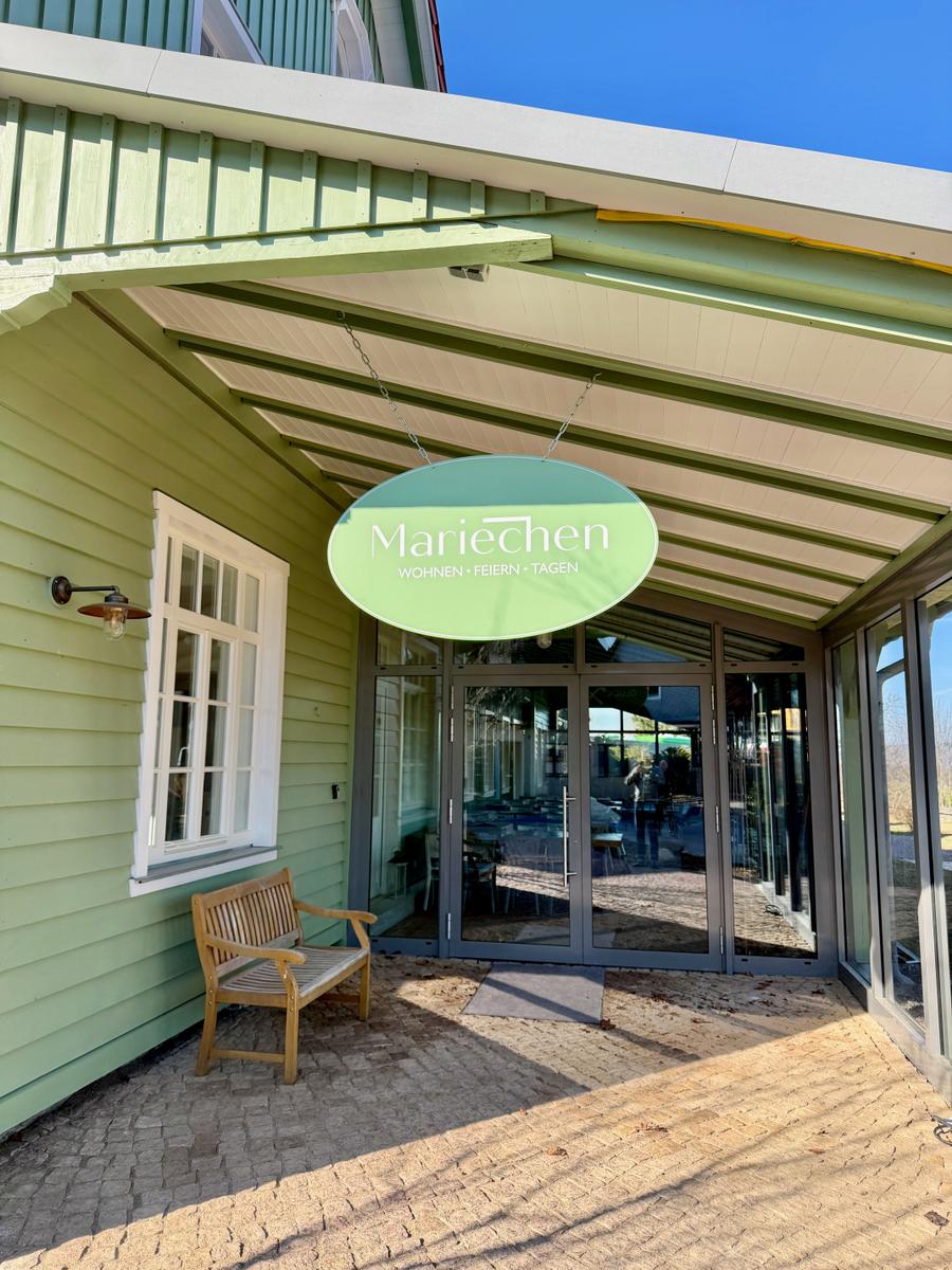 Eingangsbereich mit Schild, Glasfront und Holzbank vor grünem Haus