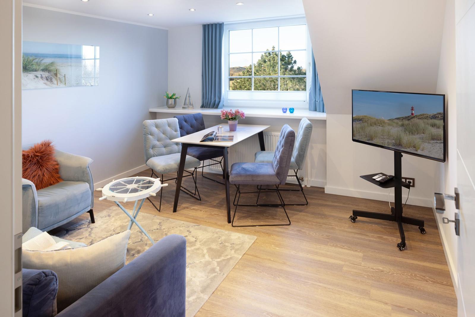 Wohn- und Essbereich mit TV, Tisch und Stühlen, Fenster mit Blick auf Grün.