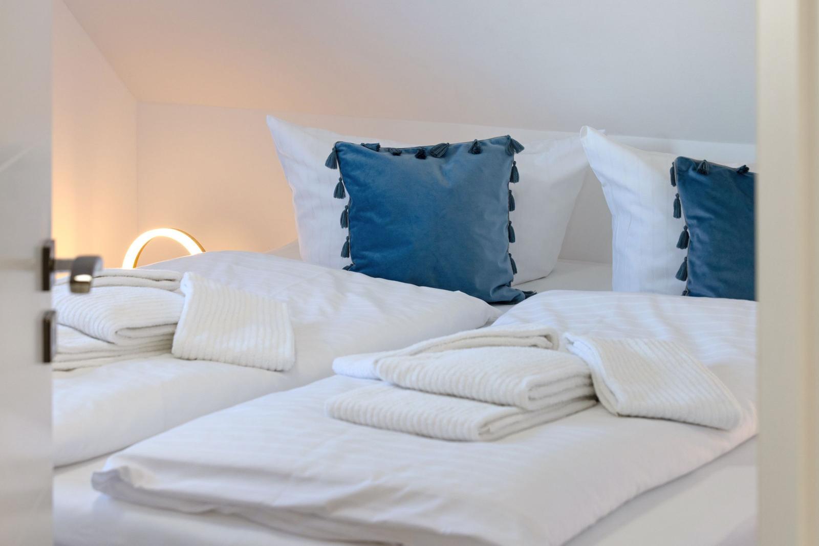 Schlafzimmer mit weißem Bett, blauen Kissen und gefalteten Handtüchern.
